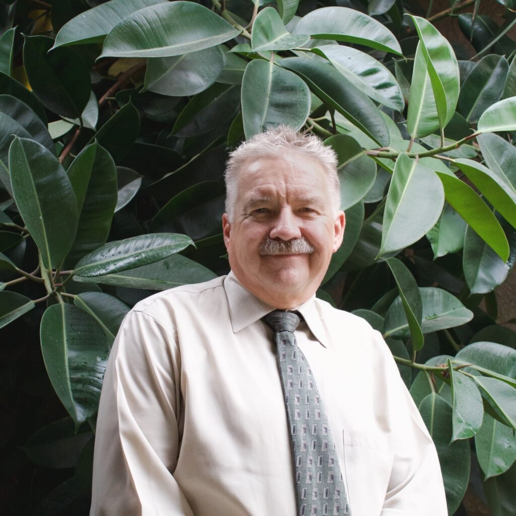 Dr. David Newsham smiling in a family-friendly dental office setting, surrounded by lush green plants, emphasizing compassionate dental care for patients of all ages.