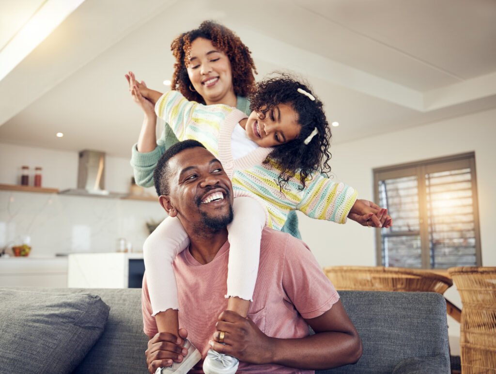 Family enjoying quality time at home, father with daughter on shoulders, smiling mother, cozy living space.