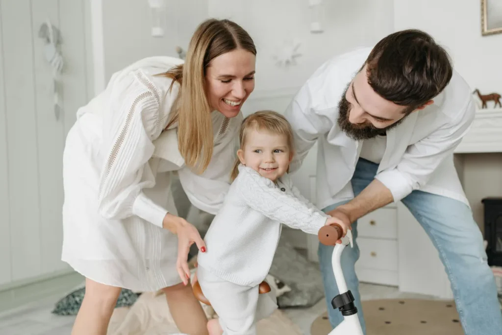 mom and dad teaching their toddler how to ride a bicycle in their home
