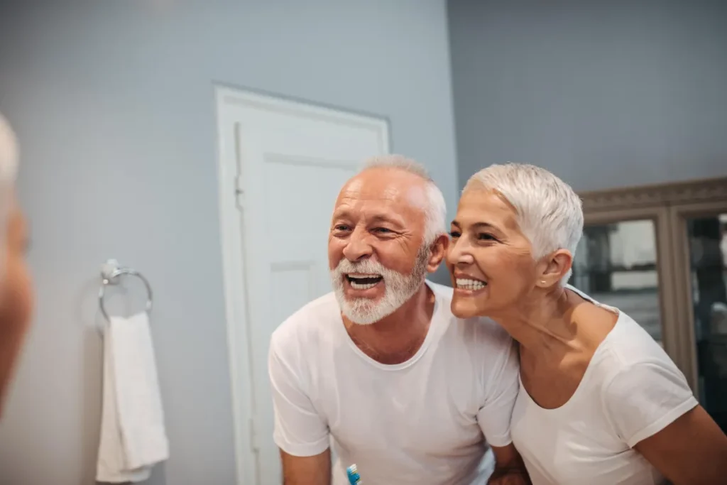 Older couple smiling in mirror admiring their smile makeover