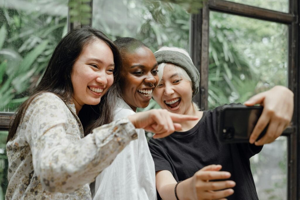 three women in college all smiling and pointing at the phone while they are taking a selfie
